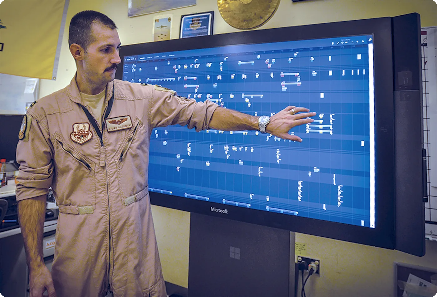 Capt. Gary Olkowski, in a flight suit, points at a large wall display showing an older version of Jigsaw's planning grid