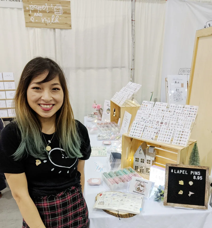 Denise smiling behind her Paper and Milk booth at a craft fair, with sticker sheets displayed on wooden stands
