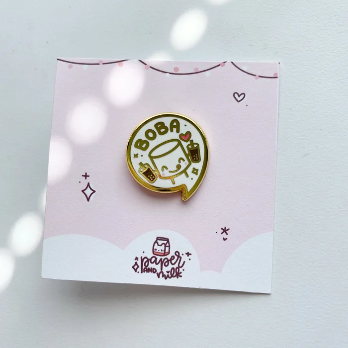 A gold enamel pin of a smiling boba cup on a pink Paper and Milk backing card